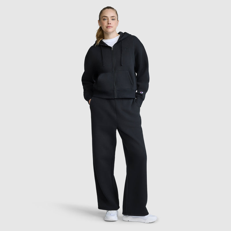 A woman stands against a plain background, wearing a black zip-up hoodie, a white shirt, white sneakers, and Champion Wide Leg Fleece Sweatpants with the C Logo. She has her hands in her pockets and looks at the camera.