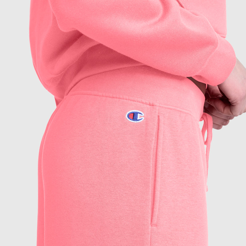 Close-up of a person wearing Champion Wide Leg Fleece Sweatpants, C Logo, in light pink. The embroidered Champion C logo is visible near the pocket of the pants. The wide leg fleece design creates a relaxed look against the plain light gray background.