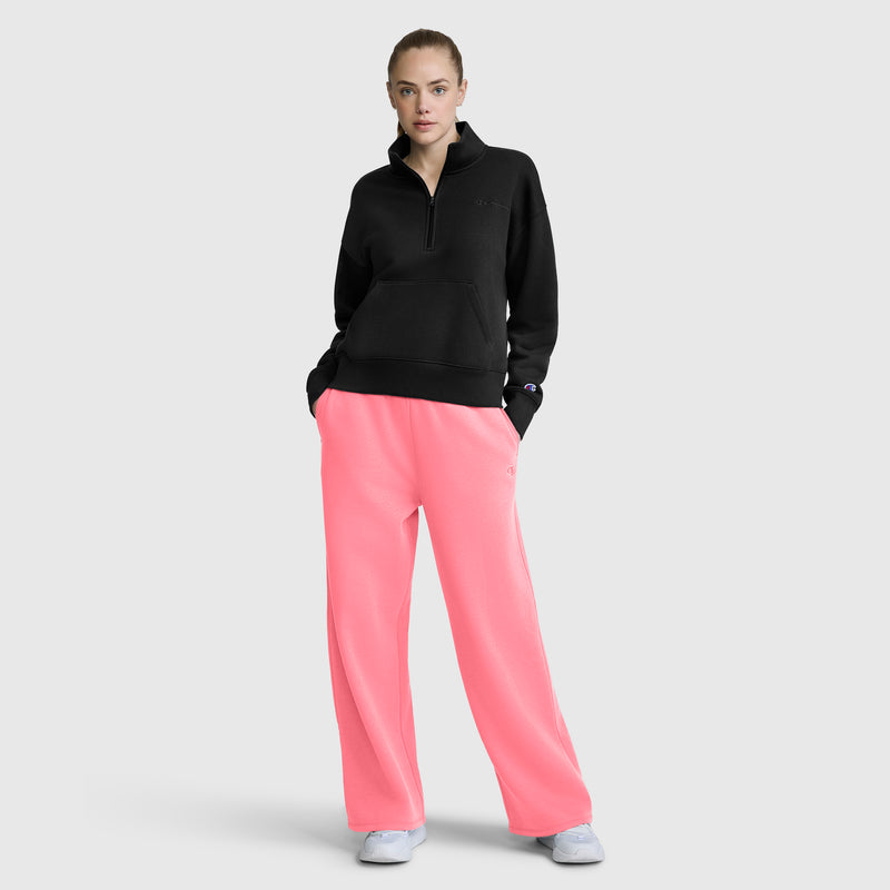 A woman stands with her hands in her pockets, wearing a black sweatshirt, Champion Wide Leg Fleece Sweatpants with C Logo in bright pink, and white sneakers against a plain light gray background.