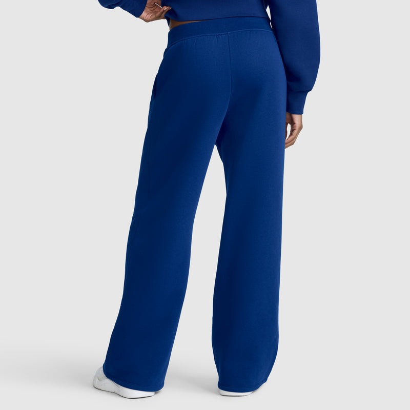 A person is shown from the back wearing blue Wide Leg Fleece Sweatpants, C Logo by Champion paired with a matching blue sweatshirt. They stand with one hand on their hip and white sneakers visible, highlighting the relaxed fit for both comfort and style.