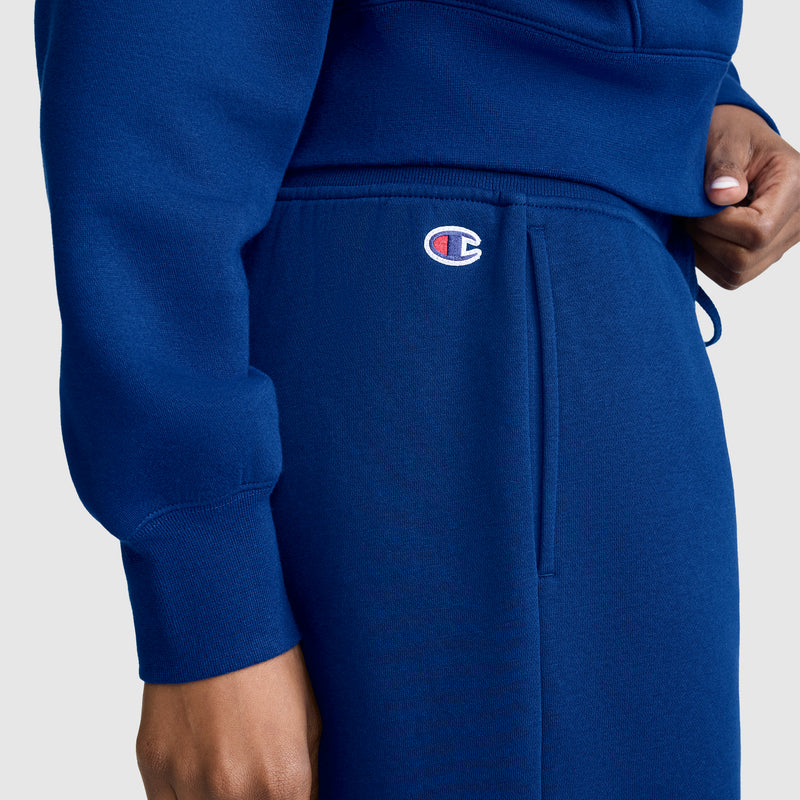 A person is wearing a blue Champion Wide Leg Fleece Sweatpants, C Logo, featuring the Champion logo above the pocket, standing against a plain light background.