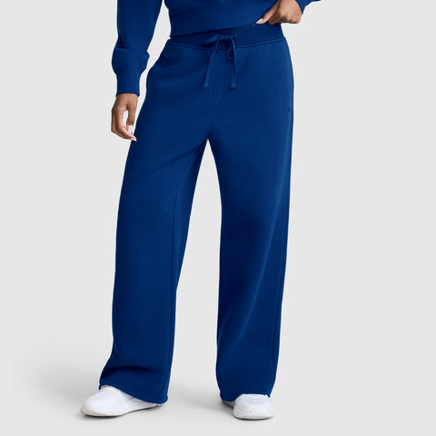 A person is seen wearing Champion Wide Leg Fleece Sweatpants, C Logo, in a loose-fitting royal blue style with a drawstring waist and white sneakers, standing against a light gray background; only the lower body is fully visible.