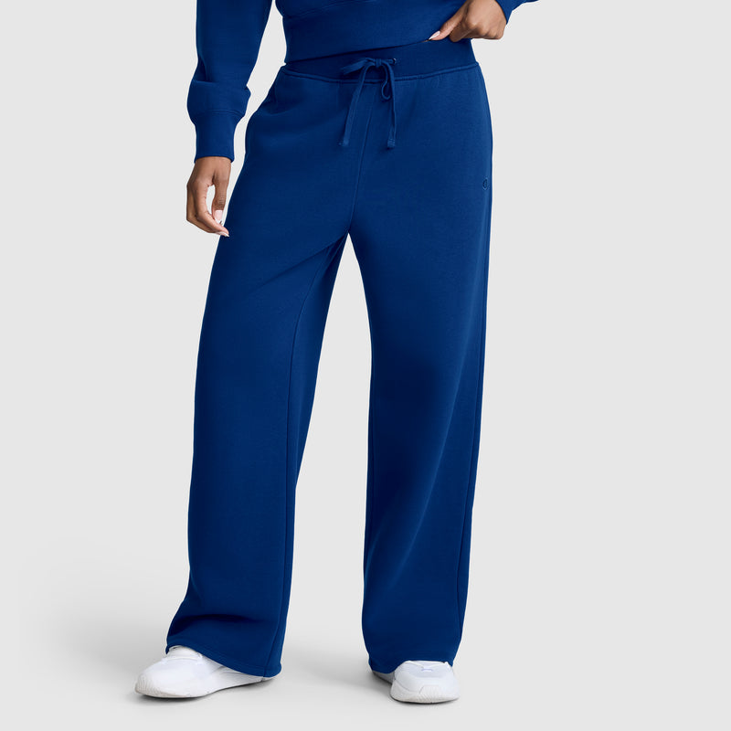 A person is seen wearing Champion Wide Leg Fleece Sweatpants, C Logo, in a loose-fitting royal blue style with a drawstring waist and white sneakers, standing against a light gray background; only the lower body is fully visible.