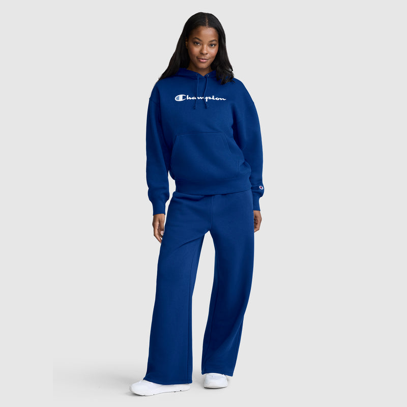 A woman with long hair poses against a plain light background, wearing white sneakers and a coordinated blue set featuring the Champion Wide Leg Fleece Sweatpants, C Logo, and a hoodie with Champion branding. One hand rests on her hip.