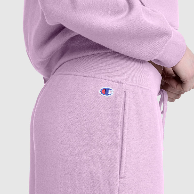 A person is wearing Champion's Wide Leg Fleece Sweatpants, C Logo, in light purple with the Champion logo visible near the waistband, paired with a matching sweatshirt and standing with hands clasped.