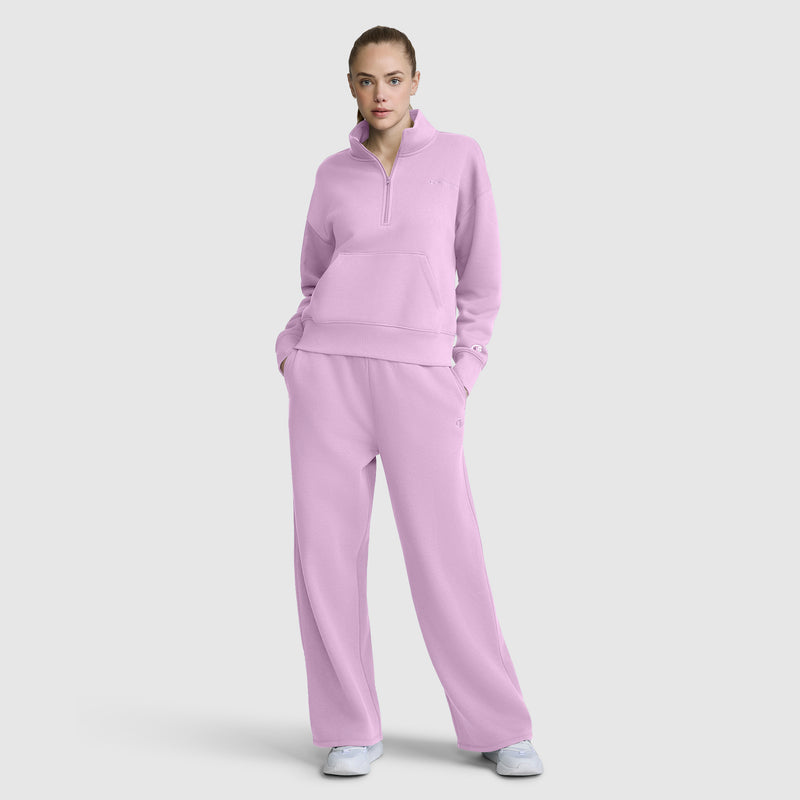 A woman stands with her hands in her pockets, wearing Champion's Wide Leg Fleece Sweatpants featuring the C Logo and a coordinating light purple quarter-zip sweatshirt, paired with white sneakers against a plain light background.