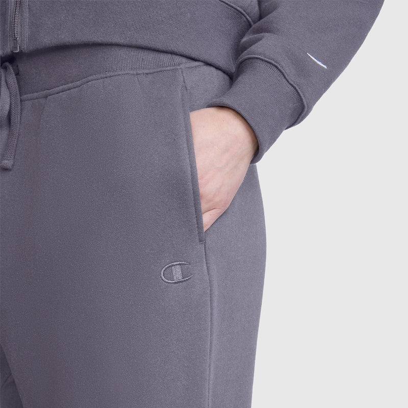 A person is wearing Champion Wide Leg Fleece Sweatpants, C Logo in grey with a matching sweatshirt, one hand in their pocket. The Champion logo is visible on the pants.