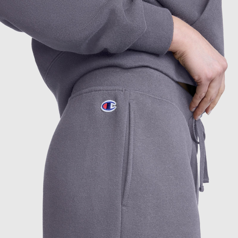 Close-up of a person wearing Champion’s Wide Leg Fleece Sweatpants, C Logo, in gray, paired with a matching sweatshirt. The pants feature a small Champion logo detail and the person has their hand resting on their hip.