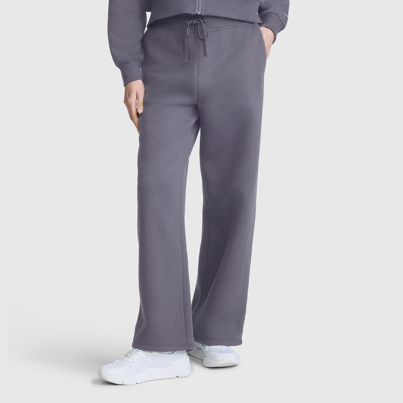 A person is shown from the waist down wearing loose-fitting Champion Wide Leg Fleece Sweatpants, C Logo, in gray, paired with white sneakers and a matching gray jacket. The modern silhouette of the Champion sweatpants stands out against a plain white background.
