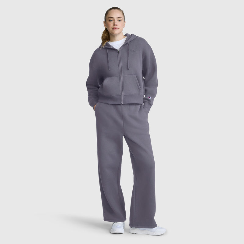 A person stands wearing Champion’s Wide Leg Fleece Sweatpants with the C Logo, styled as part of a matching gray set and paired with white sneakers. Their hands are in their pockets, and they gaze forward against a plain light background, highlighting the modern silhouette of the look.
