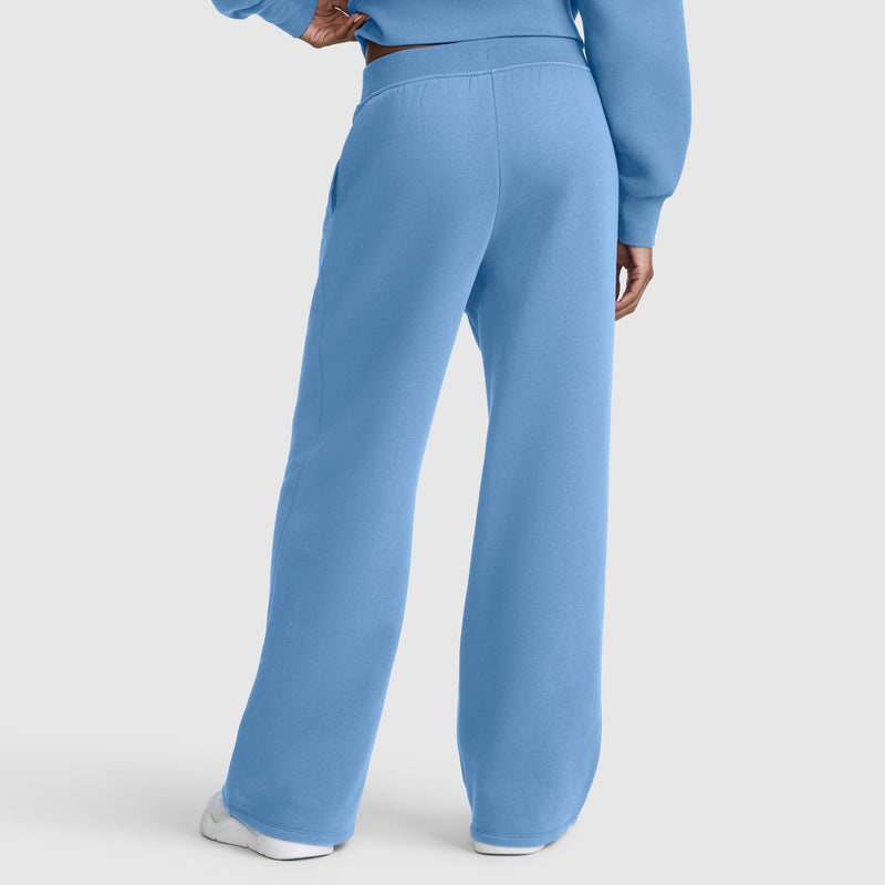 A person wearing Champion Wide Leg Fleece Sweatpants with the C Logo in light blue and a matching sweatshirt stands with one hand on their hip, facing away from the camera. White shoes are visible.