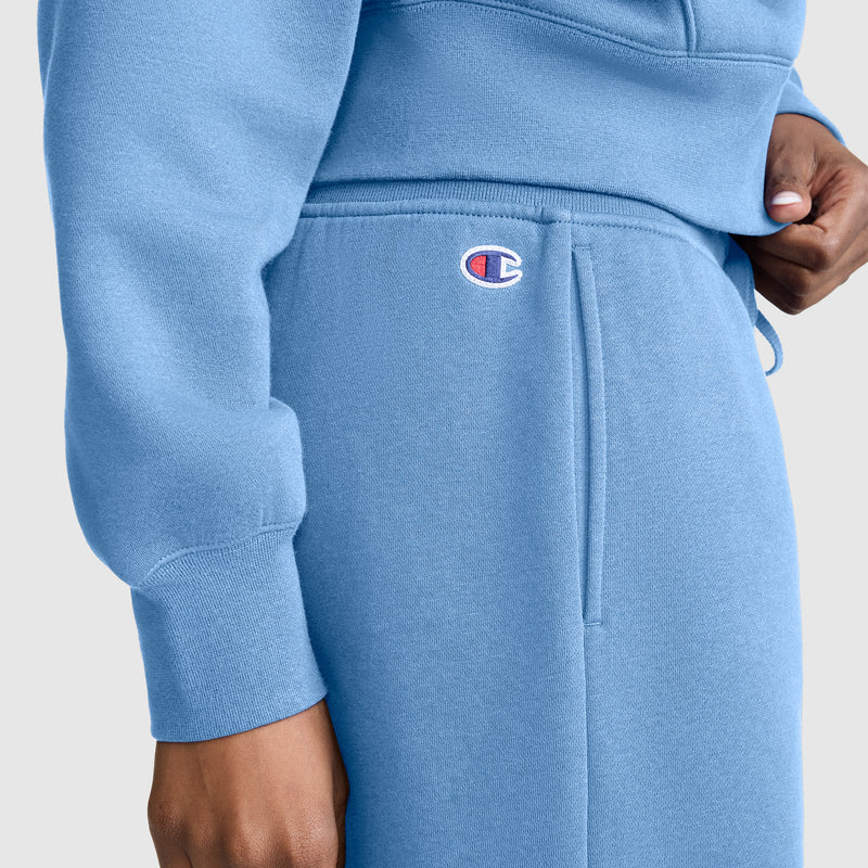 A person wears Champion Wide Leg Fleece Sweatpants with the C Logo in light blue, paired with a matching sweatshirt. Only their torso and right hand are visible, highlighting the iconic Champion logo on the wide leg sweatpants.