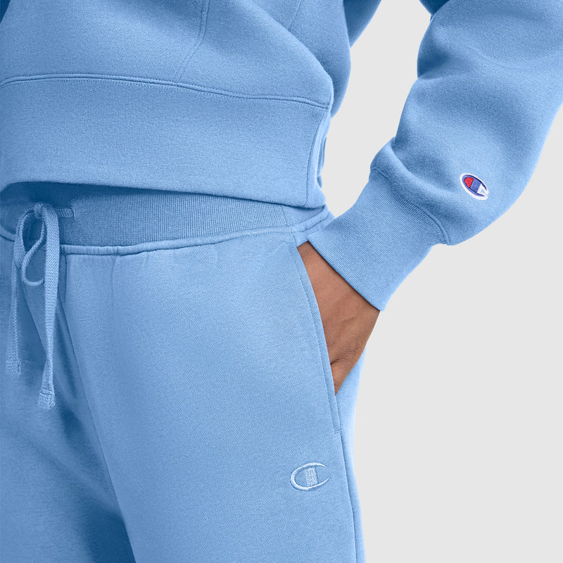 A person wearing a light blue Champion sweatshirt paired with Champion Wide Leg Fleece Sweatpants, C Logo, stands with hands in pockets. Both garments feature the Champion logo. The background is plain light gray.