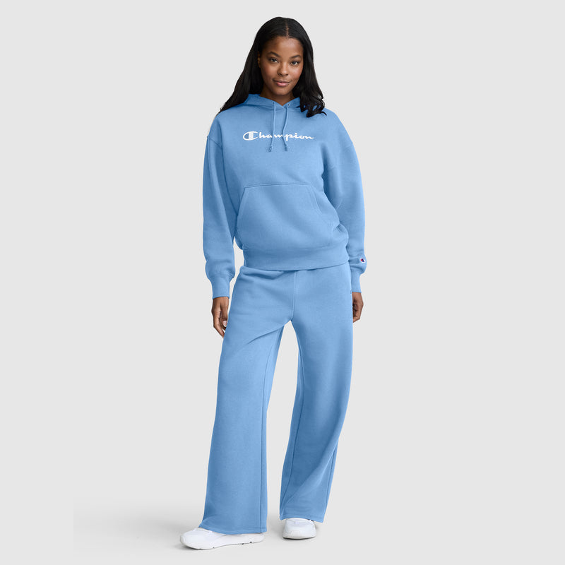 A woman stands and smiles with her right hand in her pocket, wearing a light blue Champion hoodie and matching Champion Wide Leg Fleece Sweatpants with the C Logo. She completes the look with white sneakers.