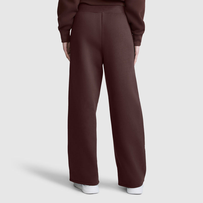 A person wearing Champion’s Wide Leg Fleece Sweatpants with a C Logo and a matching brown sweatshirt stands with their back to the camera. They are also wearing white sneakers, against a plain, light gray background.