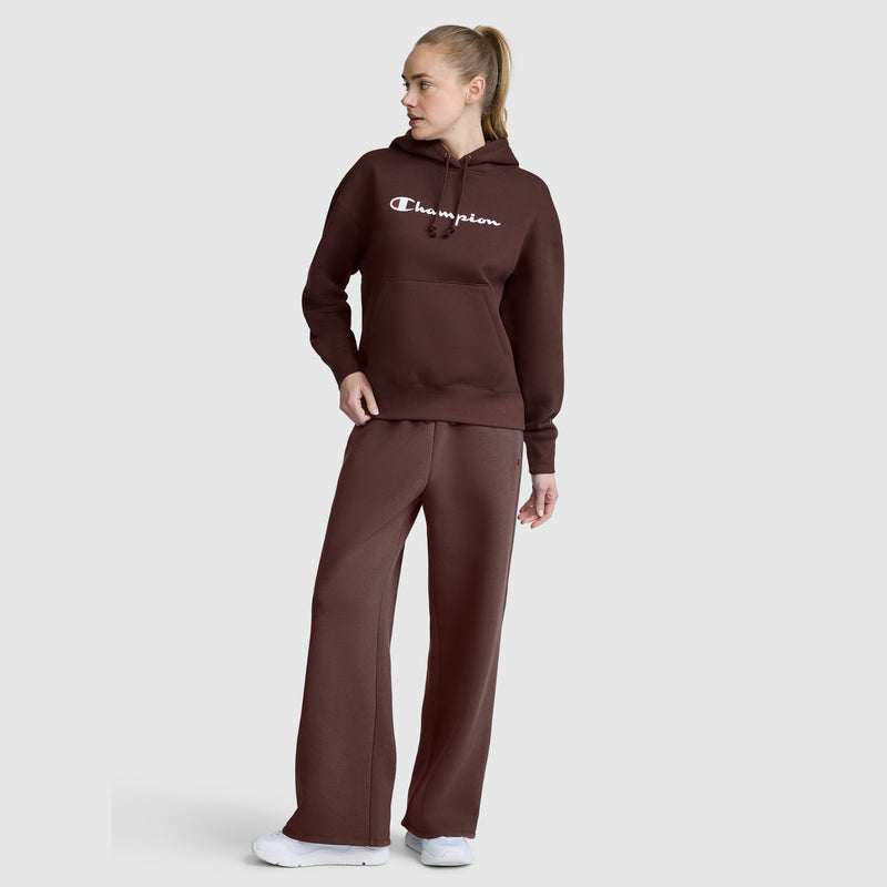A woman with blonde hair in a ponytail stands sideways against a light gray background, looking off to her left. She is wearing white sneakers and a brown Champion hoodie paired with Champion Wide Leg Fleece Sweatpants, C Logo.