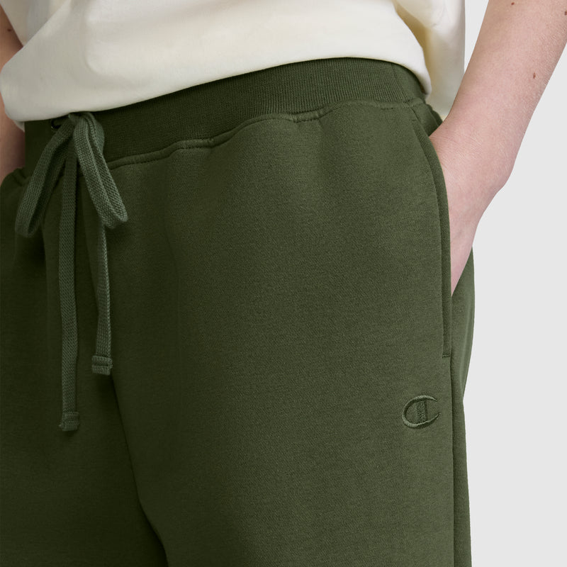 A person wearing Champion’s Wide Leg Fleece Sweatpants with the signature C Logo in olive green, featuring a drawstring waist and a relaxed wide leg fleece style; one hand in the pocket and a white shirt partially visible.