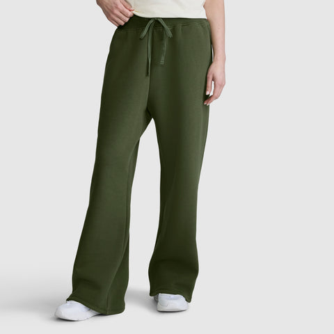 A person is wearing Champion Wide Leg Fleece Sweatpants, C Logo in olive green, which feature a high-waisted design with an elastic drawstring waistband and wide legs. The sweatpants are styled with white sneakers and photographed against a plain light gray background.