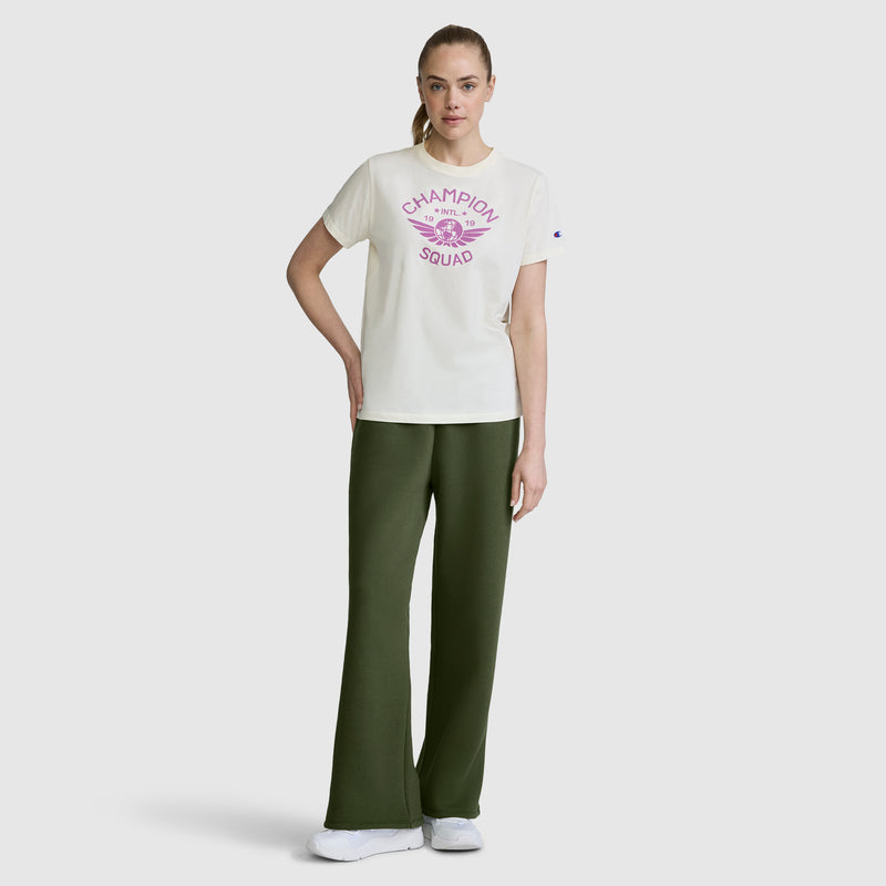 A woman stands against a plain light background, wearing a white Champion Squad t-shirt with pink lettering paired with Champion Wide Leg Fleece Sweatpants featuring the C Logo in dark green, and white sneakers. One hand rests on her hip while her other arm is relaxed.