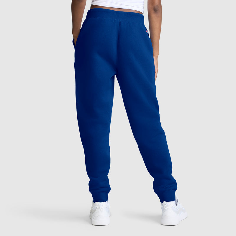 A person wearing Champion Powerblend Joggers with the Classic Script Logo in royal blue, styled with a white crop top and white sneakers, stands with hands in pockets against a plain light background. Only the lower body is visible.