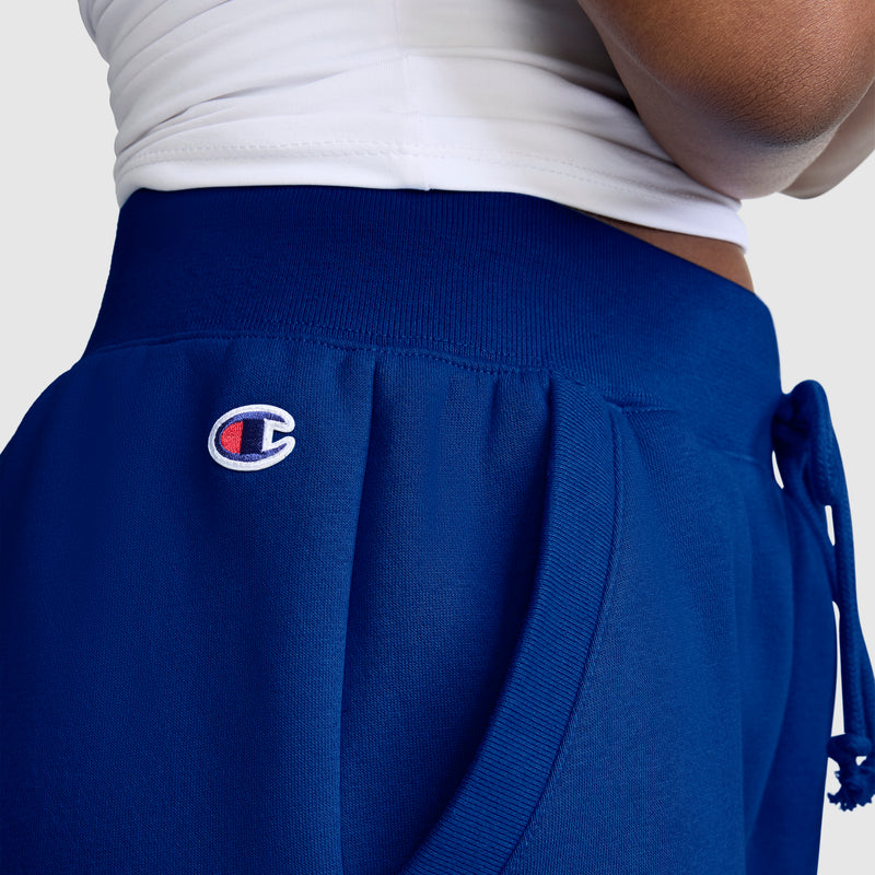 Close-up of a person wearing blue Champion Powerblend Joggers, Classic Script Logo, featuring a white drawstring and paired with a white top. The joggers display the Champion logo on the left hip and offer both comfort and style, making them an essential choice among women’s joggers.