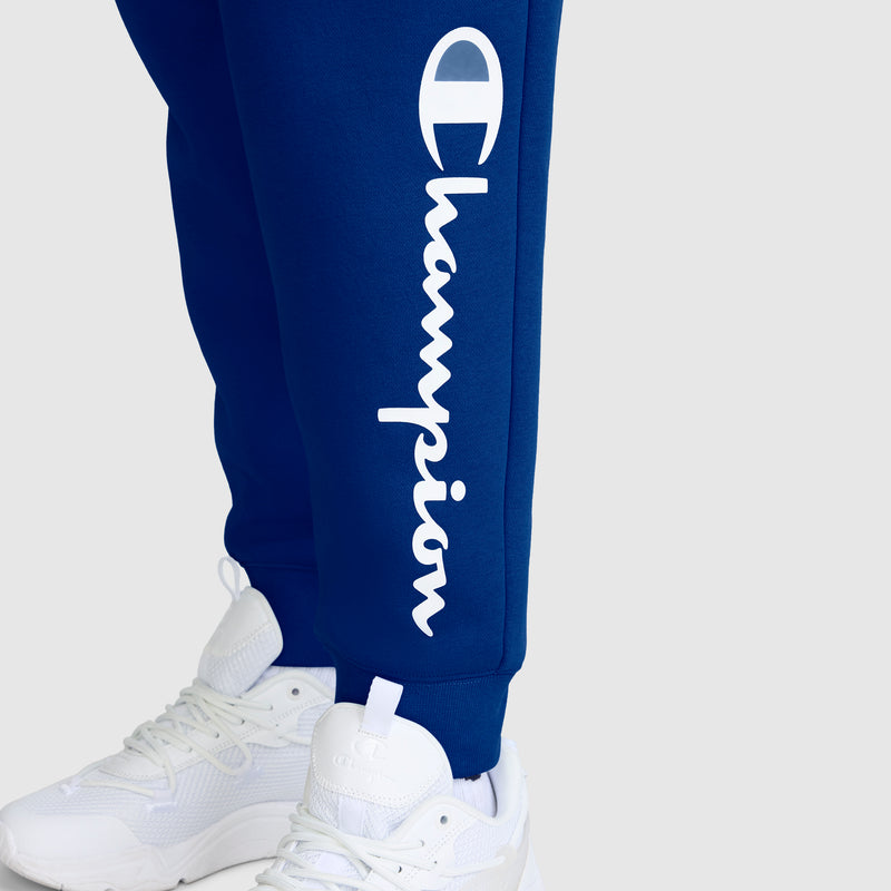 A person is wearing blue Champion Powerblend Joggers with the Classic Script Logo printed vertically in white on the left leg, paired with white sneakers. These cozy joggers for women from Champion stand out with their signature branding.