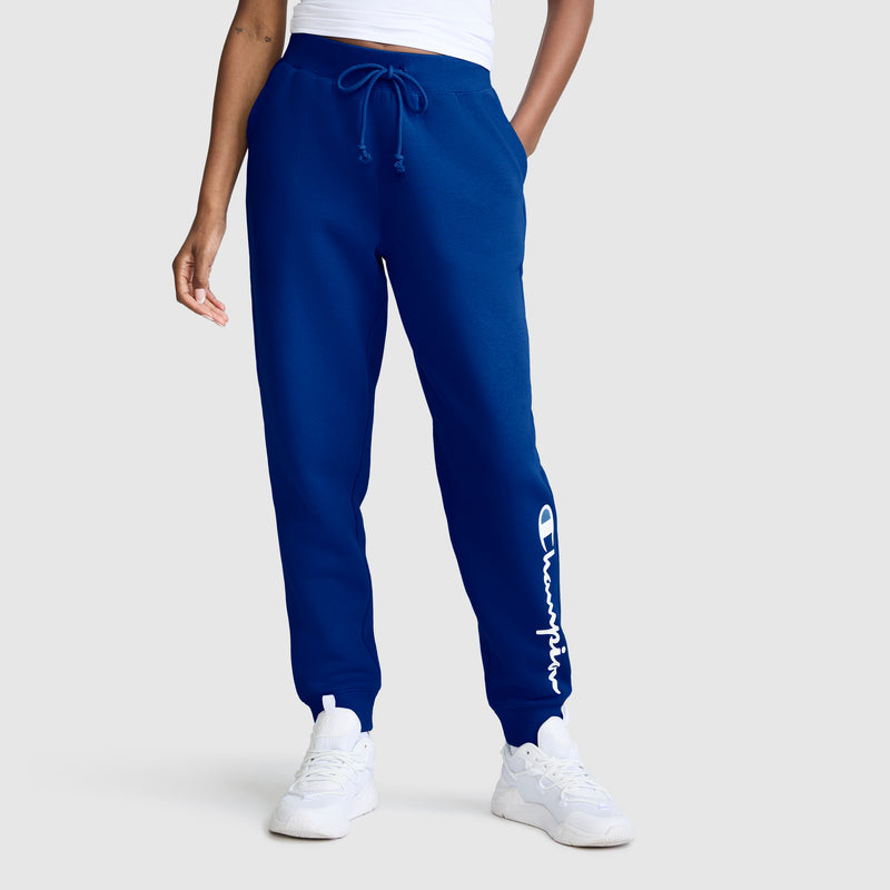 A person wears the Champion Powerblend Joggers with the Classic Script Logo on the left leg, styled with a white top and white sneakers, standing against a plain light background.