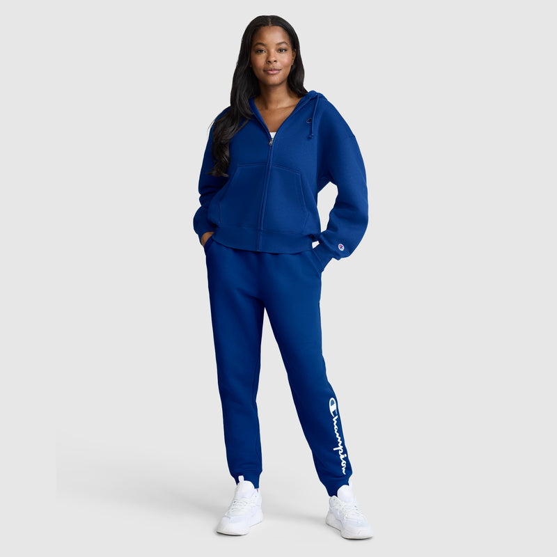 A woman stands smiling with her hands in the pockets of her matching blue Champion Powerblend Joggers with Classic Script Logo and a zip-up hoodie, paired with white sneakers, against a plain light background.