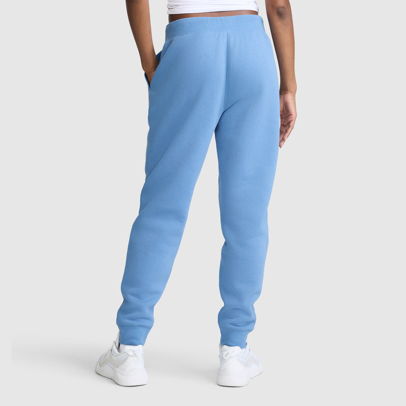 A person is shown from the back against a plain light gray background, wearing Champion Powerblend Joggers with the Classic Script Logo in light blue, paired with a white cropped top and white sneakers.