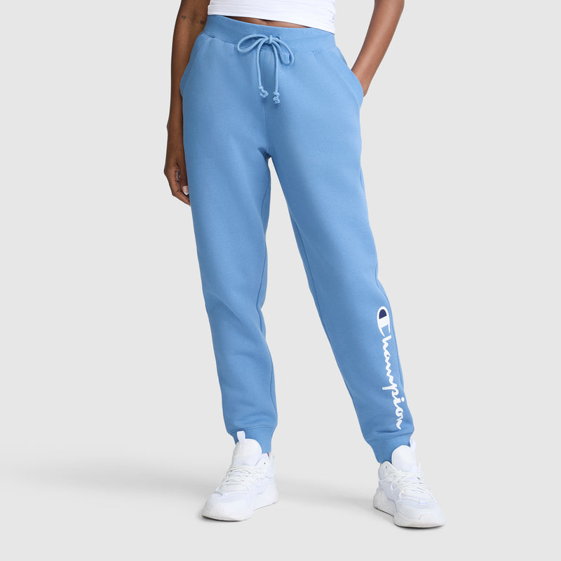 A person is wearing Champion Powerblend Joggers, Classic Script Logo, in light blue—featuring the signature Champion logo on the left leg. They’ve paired the fleece joggers with white sneakers and a white top, standing against a plain light background. These comfortable joggers for women offer a blend of style and ease, making them an ideal everyday choice.
