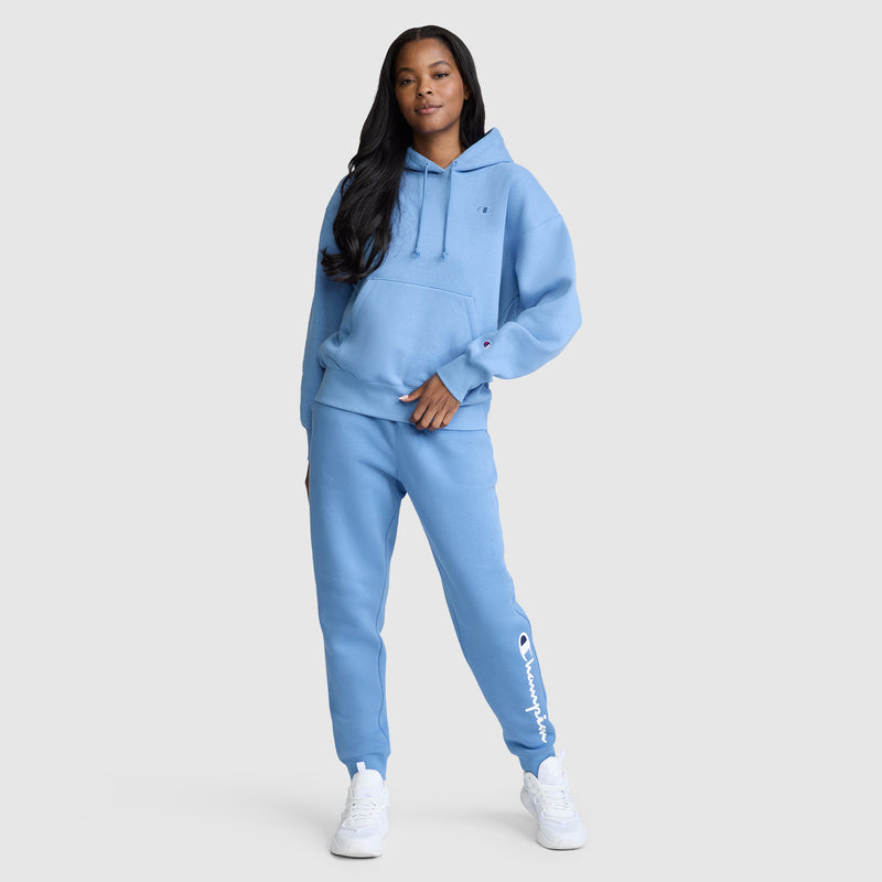 A woman stands confidently with one hand in her pocket, wearing the Champion Powerblend Joggers with the Classic Script Logo in light blue and a matching hoodie. She completes her look with white sneakers and has long, dark hair. The background is plain and light gray.