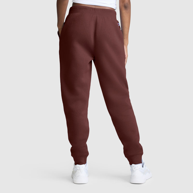 A person stands with their back to the camera against a plain light gray background, wearing the Champion Powerblend Joggers, Classic Script Logo in maroon, paired with white sneakers and a white top—perfect for anyone looking for comfortable and stylish women’s joggers.