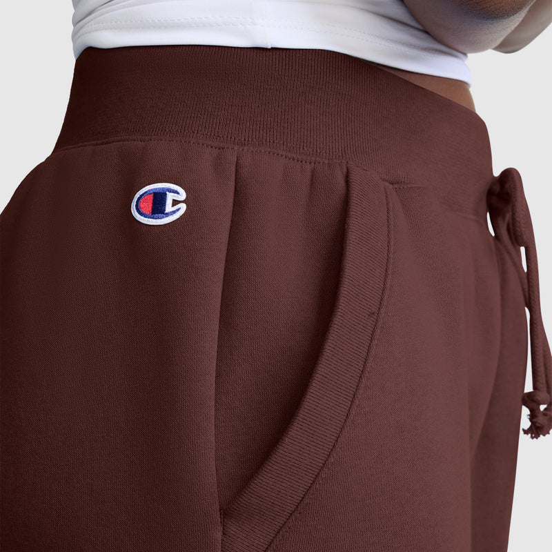 Close-up of a person wearing maroon Champion Powerblend Joggers, Classic Script Logo, featuring the embroidered Champion logo near the pocket and a white waistband of a top visible at the top of the image. These comfortable joggers from Champion are perfect for women seeking both style and coziness.