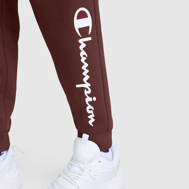 A person wearing maroon Champion Powerblend Joggers with the Classic Script Logo and white athletic sneakers, standing on a light gray background.