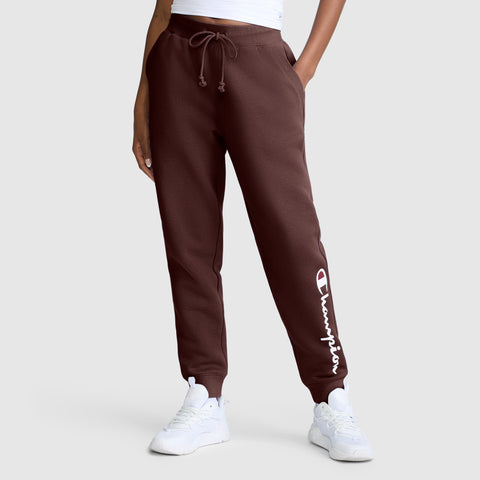 A person wearing Champion Powerblend Joggers with the classic script logo in brown, paired with white sneakers and a white top, stands against a light gray background.