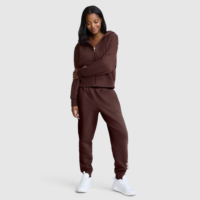 A woman stands smiling against a plain light gray background, wearing Champion Powerblend Joggers with the Classic Script Logo, a matching brown zip-up hoodie, and white sneakers.