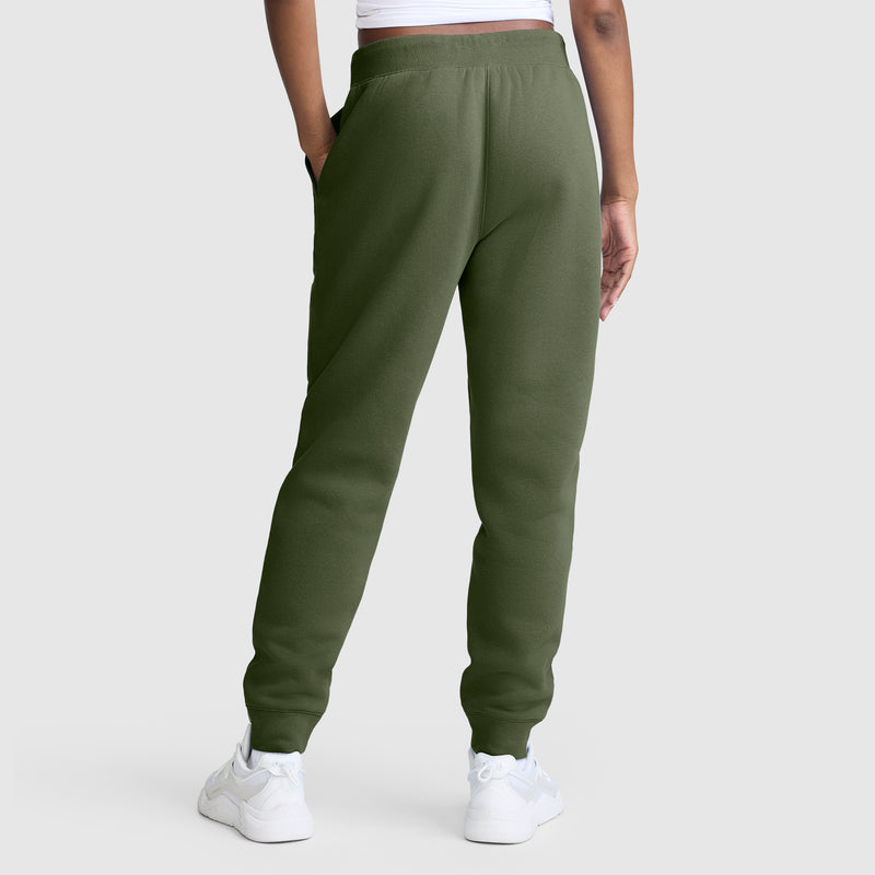 A person is shown from the back, standing against a plain light gray background, wearing Champion Powerblend Joggers with the Classic Script Logo in olive green and white sneakers.