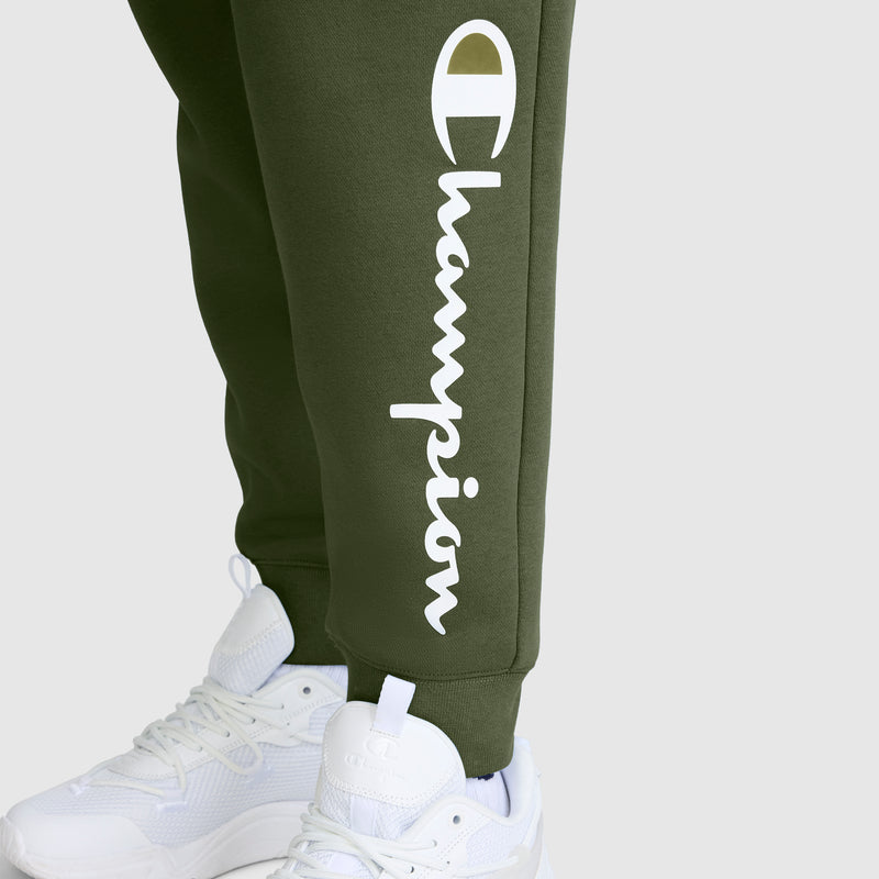 Model wears white sneakers and Champion’s Powerblend Joggers with the Classic Script Logo in olive green, featuring "Champion" boldly printed in large white letters down the leg. These comfortable joggers are perfect for everyday wear. The background is a plain, light grey.