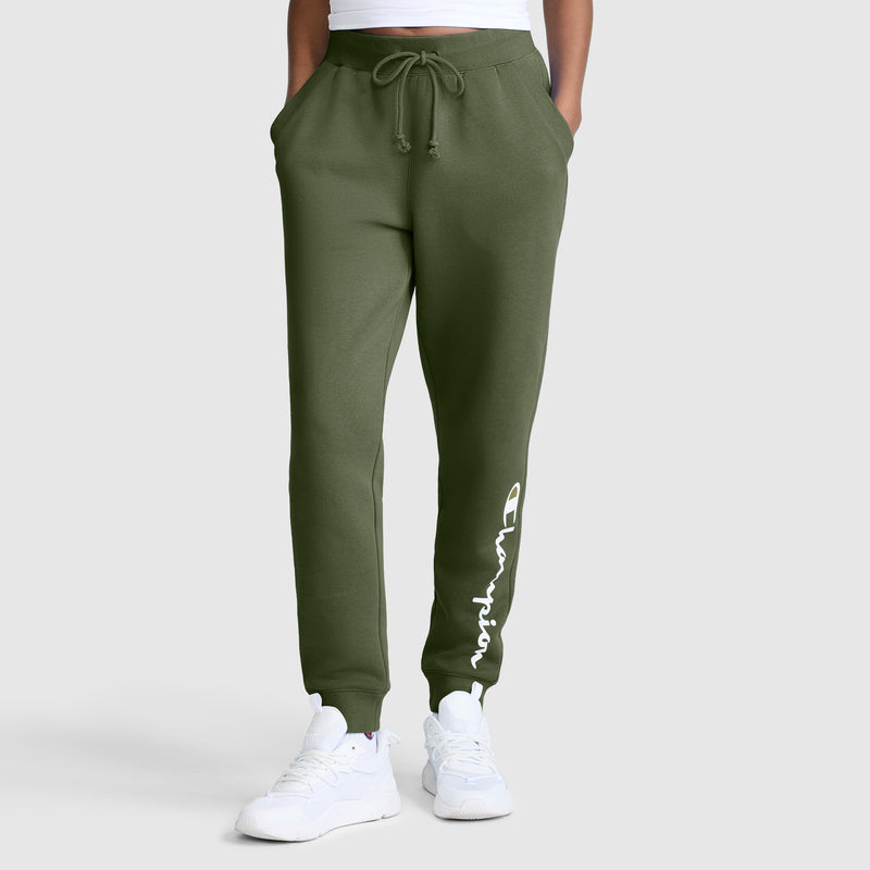 A person wearing Champion Powerblend Joggers for women in olive green, featuring the classic script logo in white on the left leg, paired with white sneakers and a white crop top. Their hands are in the joggers' pockets, set against a light gray background.