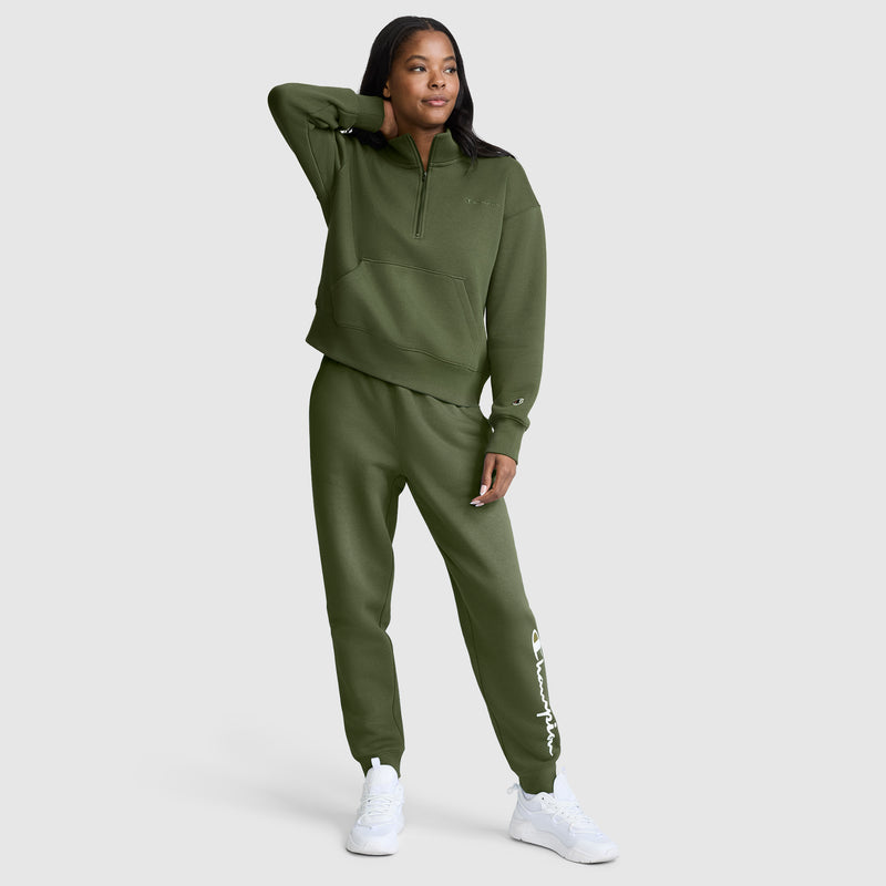 A woman in an olive green tracksuit with white sneakers stands with one hand behind her head and the other by her side, posing against a plain light gray background. Her comfortable Champion Powerblend Joggers, featuring the classic script logo, add both style and ease to her relaxed look.