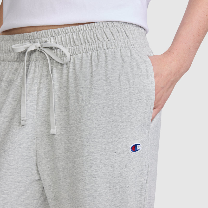 A person is wearing Champion Lightweight Lounge Pants, C Logo, in light gray with a drawstring and their right hand in the pocket. The iconic Champion C logo appears on the left leg, showcasing classic Champion quality. Crafted from soft jersey cotton, these pants are lightweight and designed for everyday comfort.