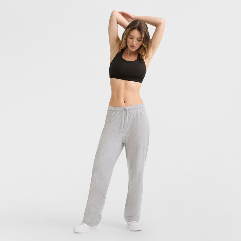 A woman wears Champion Lightweight Lounge Pants, C Logo, with a black sports bra and white sneakers as she stretches with arms raised against a plain white background.