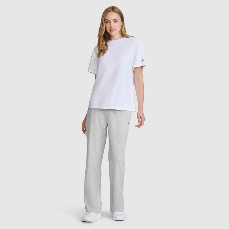 A woman stands against a plain background, wearing a white short-sleeve t-shirt, Champion Lightweight Lounge Pants with the C Logo in light gray, and white sneakers. She has long, wavy blonde hair and a neutral expression.