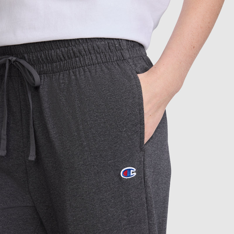 Wearing the Champion Lightweight Lounge Pants, C Logo in dark gray—renowned for their Champion quality and ultra-soft comfort—a person stands with one hand in the pocket. Only the lower half of a white shirt and a single arm are visible.