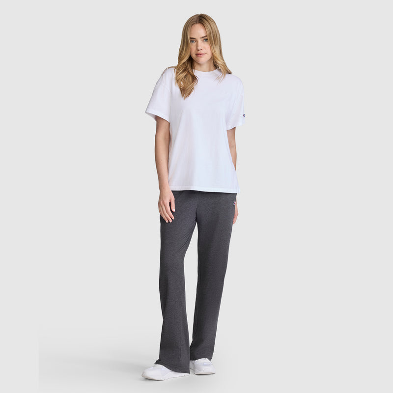 A woman with long blonde hair stands facing forward, wearing a white t-shirt, Champion Lightweight Lounge Pants with C Logo in dark gray, and white sneakers against a plain light gray background.