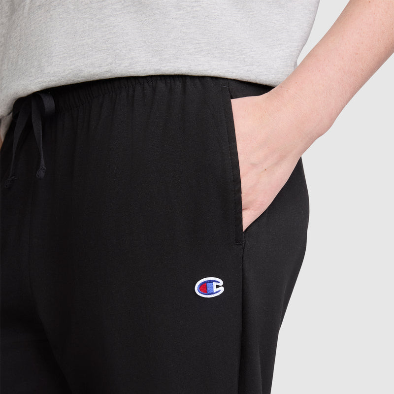 A person wearing a light gray shirt and black Champion Lightweight Lounge Pants, C Logo, with one hand in their pocket. The Champion logo is visible on the jersey cotton pants.