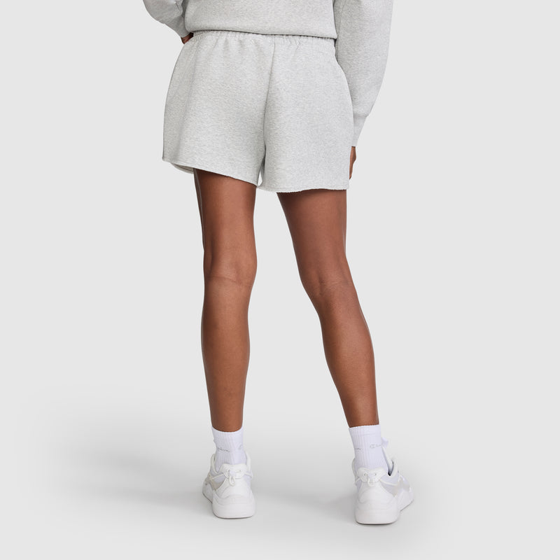 A person shown from the back wears light gray Champion 3" Powerblend Shorts, C Logo for women, a matching sweatshirt, white socks, and white sneakers. The background is plain white.