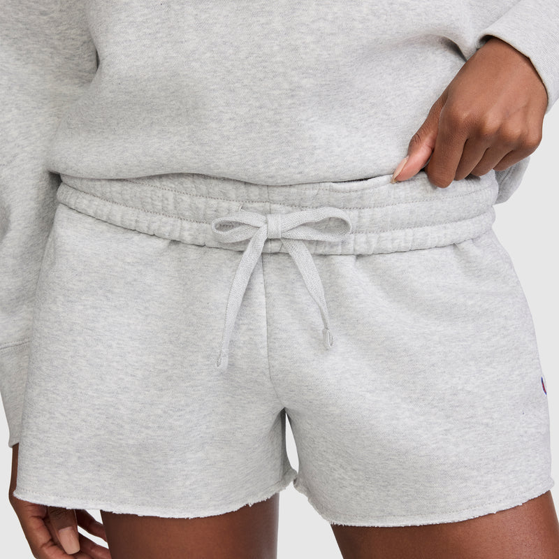 A person wearing Champion 3" Powerblend Shorts, featuring light gray fleece fabric with a drawstring waistband and the signature C Logo, paired with a matching sweatshirt. Only the lower torso and hand adjusting the waistband are visible.