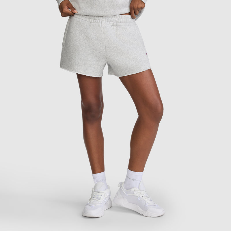 A person wearing Champion 3" Powerblend Shorts, C Logo, in light gray is shown standing with white socks and white sneakers against a plain light background. The image is cropped to display only the lower torso and legs.