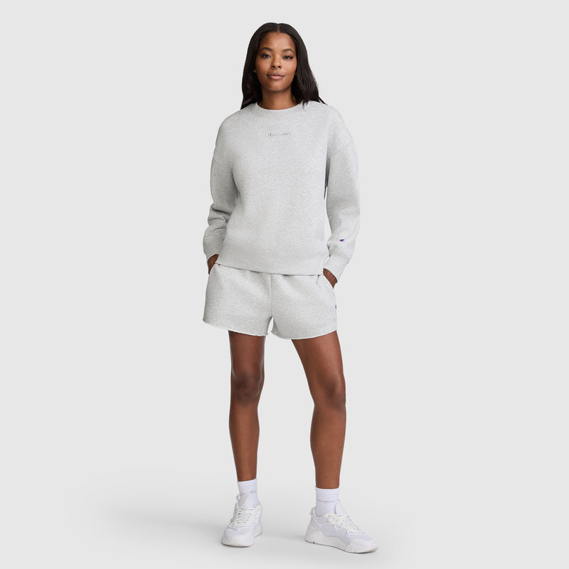 A woman stands facing forward with her hands in the pockets of her light gray sweatshirt and matching Champion 3" Powerblend Shorts, C Logo. She completes the look with white sneakers and white socks, set against a plain light gray background.